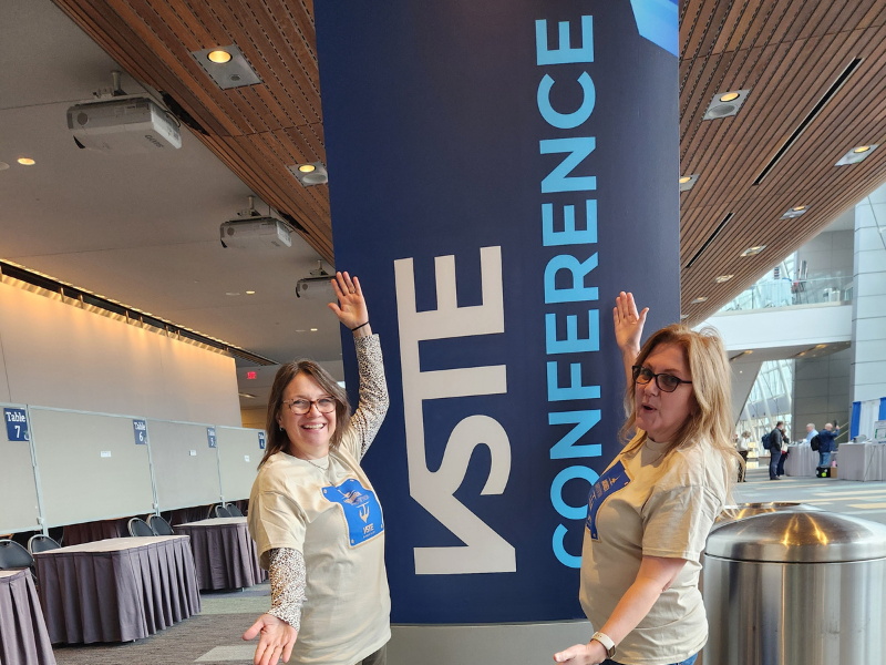 Two VSTE volunteers pointing at the VSTE Conference sign as a sign of welcome